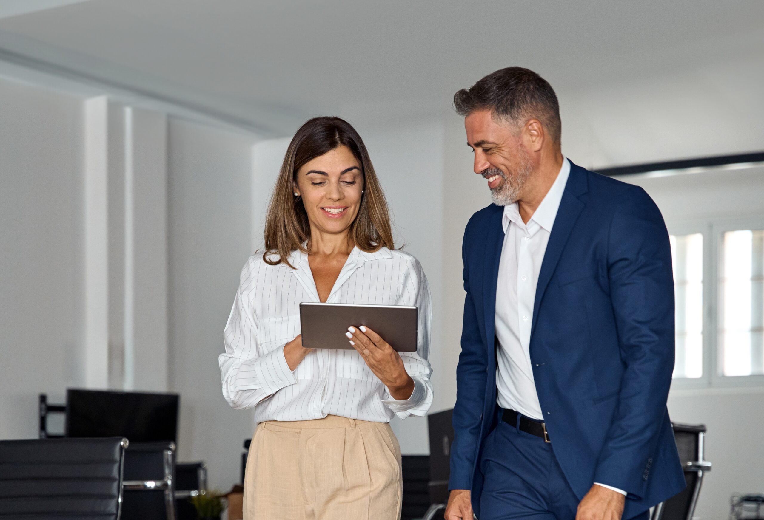 Full body vertical portrait of walking mature Latin businessman and European businesswoman discussing project on tablet in office. Two diverse partners of professional business people work together.
